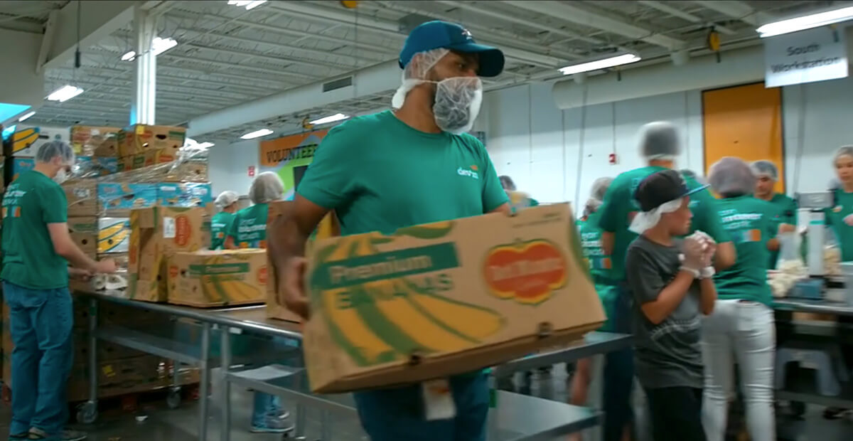 Regional food bank volunteer carrying a box of bananas while other volunteers help pack boxes.