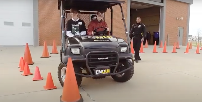 Two people in an ATV driving through a test course while an officer looks on.