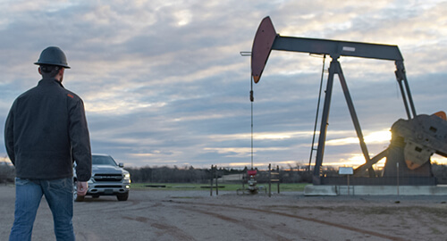 Cloudy gray sky with sun setting, pump jack with a Ford truck parked nearby and a worker with hard hat walking toward the pump jack