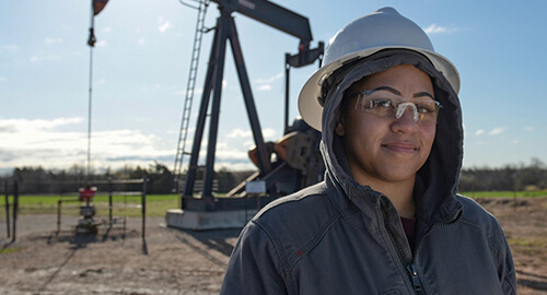 Female worker wearing a dark hoodie, white hard hat and clear goggles standing in front of a pump jack