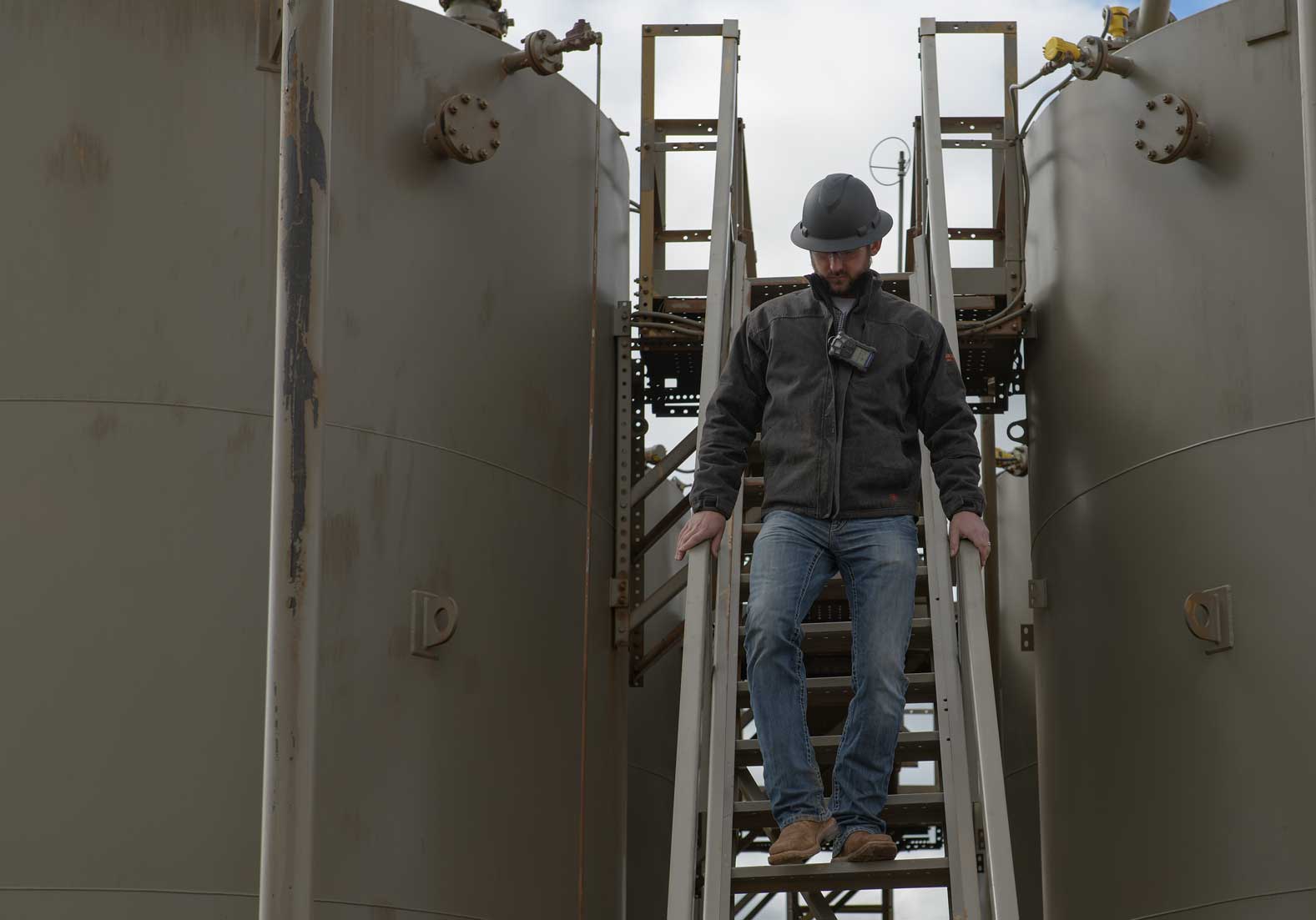 Male worker dressed in jeans, dark jacket, and black hard hat descending the stairs between two large metal tanks