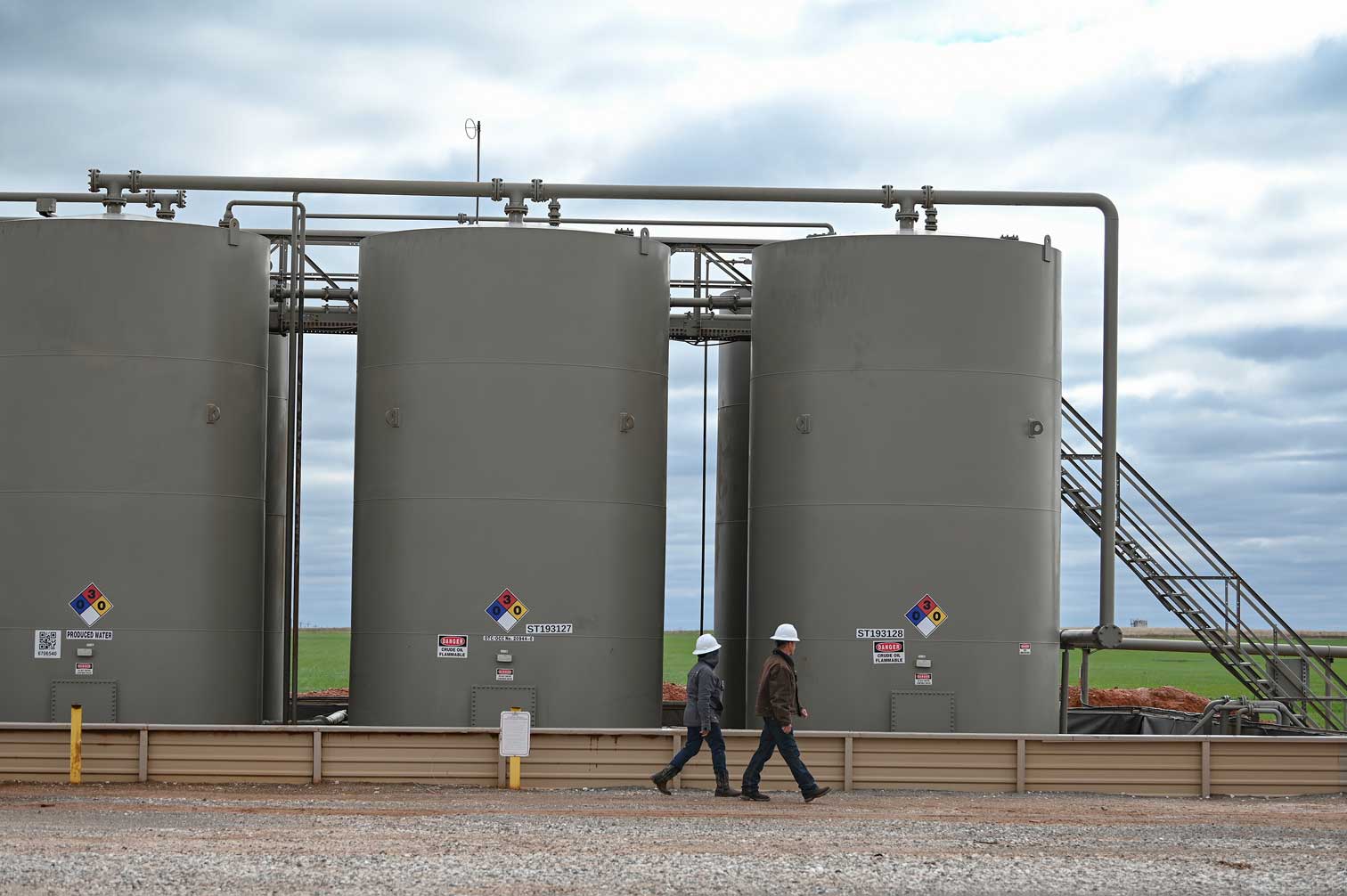 Two workers wearing jeans, dark jackets, and white hard hats while walking in front of three large metal tanks