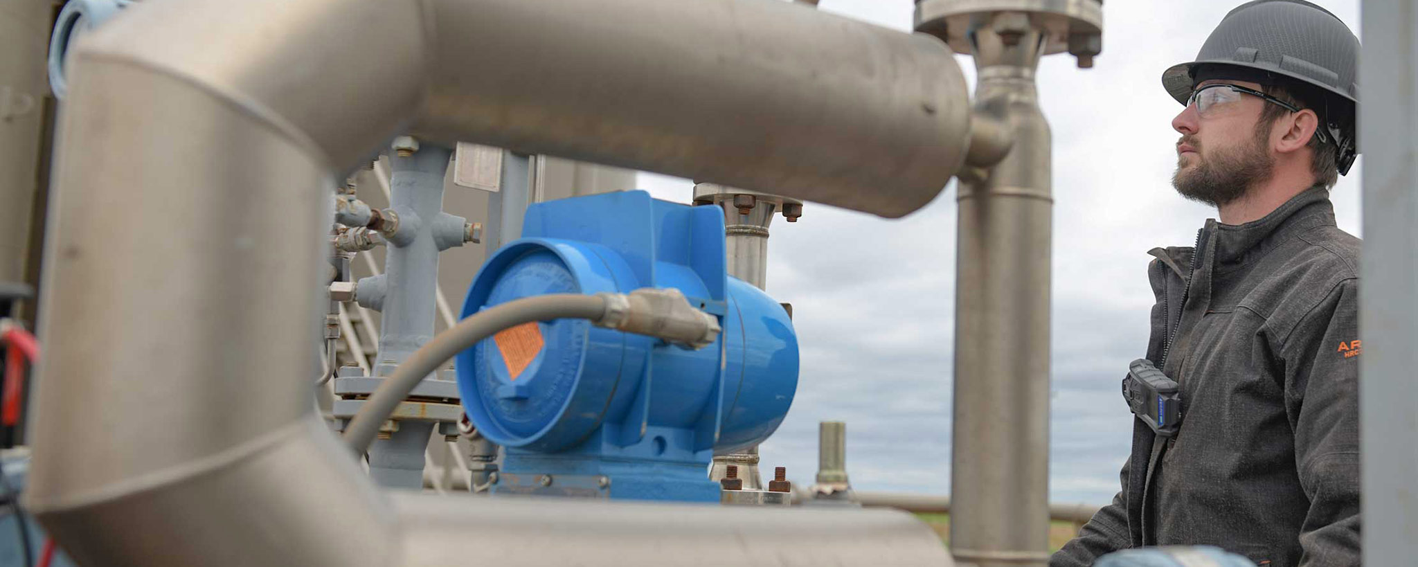 Man with beard, goggles, hard hat and black jacket looking at pump machinery