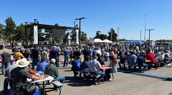 Group of attendees seated outside at tables in front of a stage